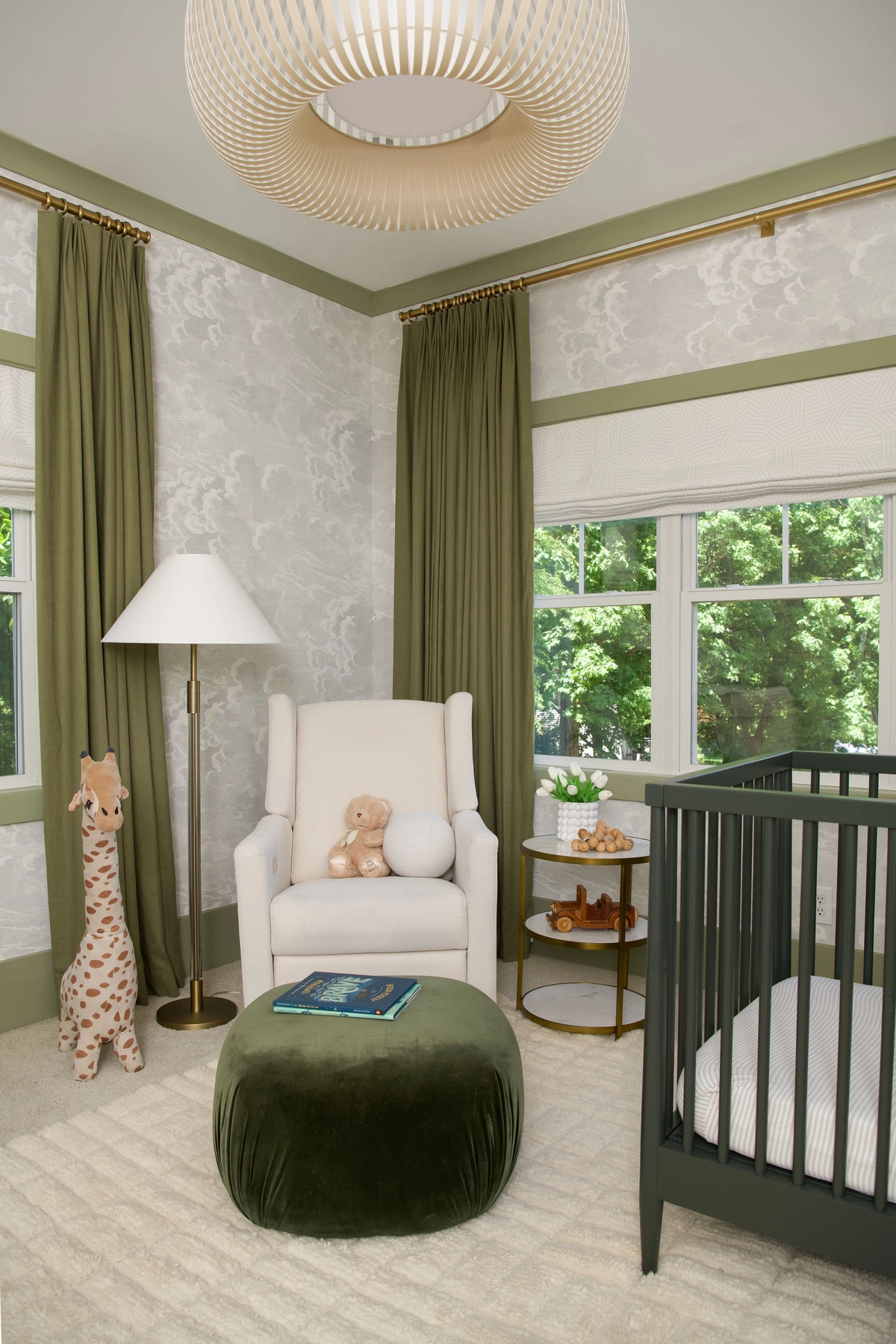 Green nursery corner with a cream glider chair, olive velvet pouf with a storybook, brass floor lamp, stuffed giraffe, cloud wallpaper, dark green crib, and slatted pendant light
