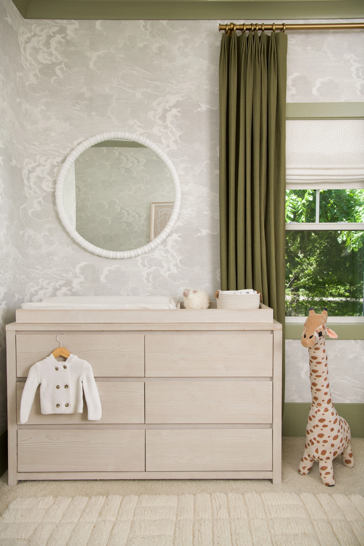 Nursery vignette with a light wood six-drawer dresser topped with a changing pad, a round white beaded mirror on cloud-motif wallpaper, olive green drapes, and a plush giraffe toy
