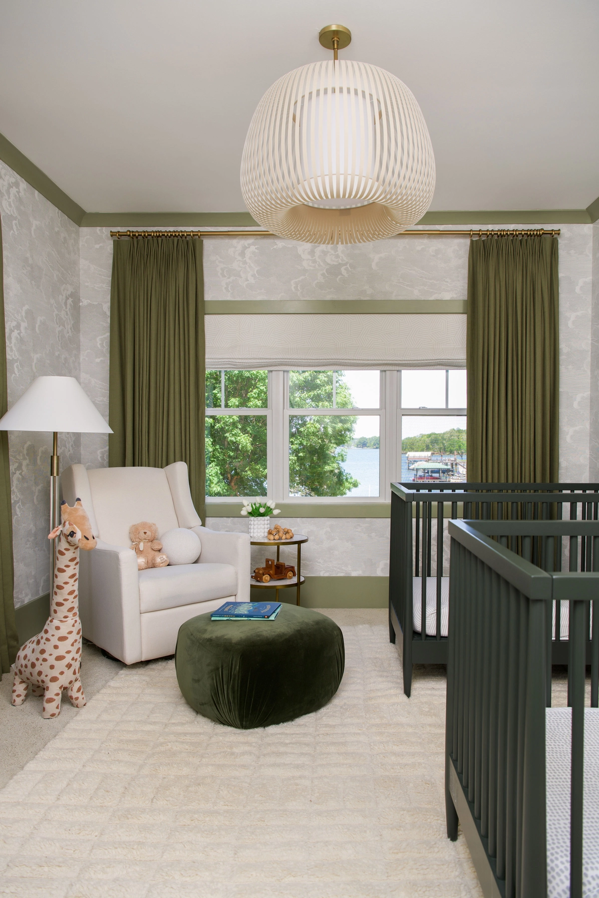 Nursery seating area with a cream upholstered glider, a dark green velvet round pouf, a brass floor lamp, olive green curtains, cloud-patterned wallpaper, and lake views