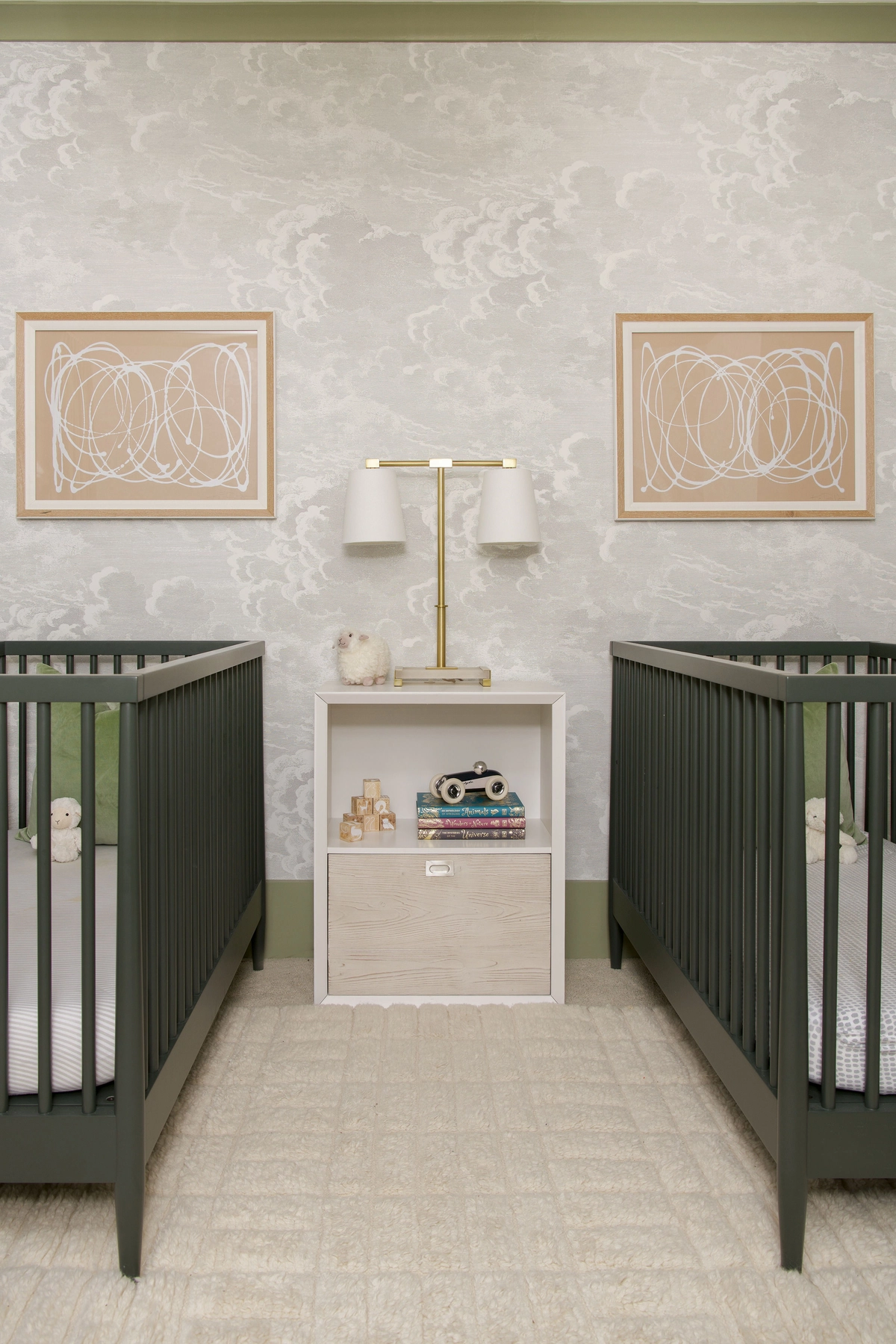 Twin nursery with two dark green cribs flanking a light wood nightstand with a brass double-arm lamp, abstract art prints above, and soft cloud-patterned wallpaper