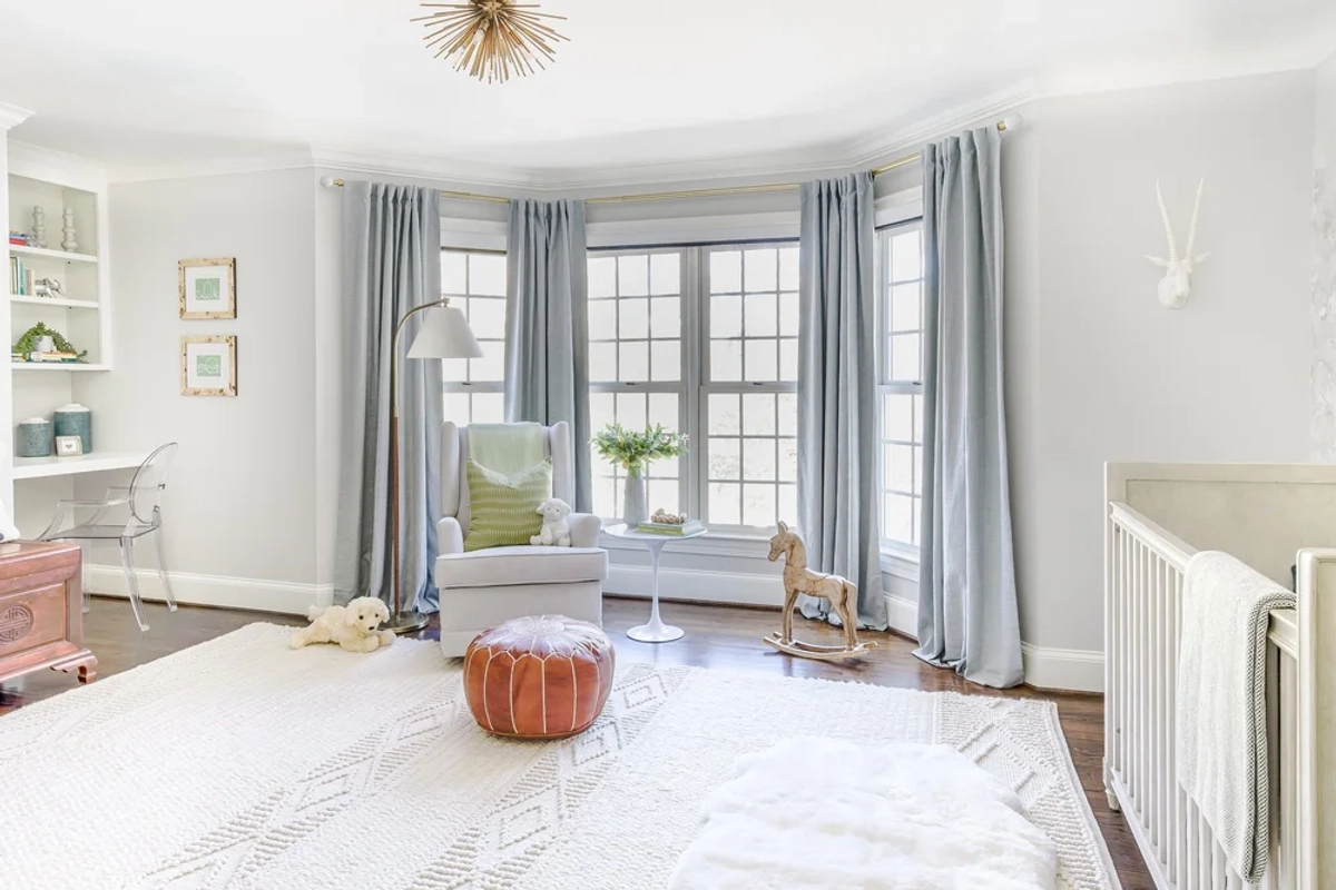 Bright nursery with a bay window dressed in soft blue curtains, a white upholstered glider chair, a cognac leather pouf, and floating shelves