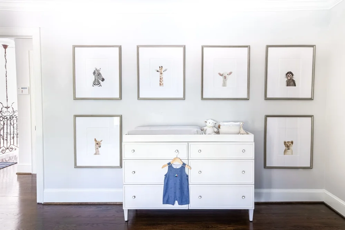 Nursery wall with a white dresser used as a changing table, a gallery wall of six framed baby animal illustrations, and a small hanging outfit