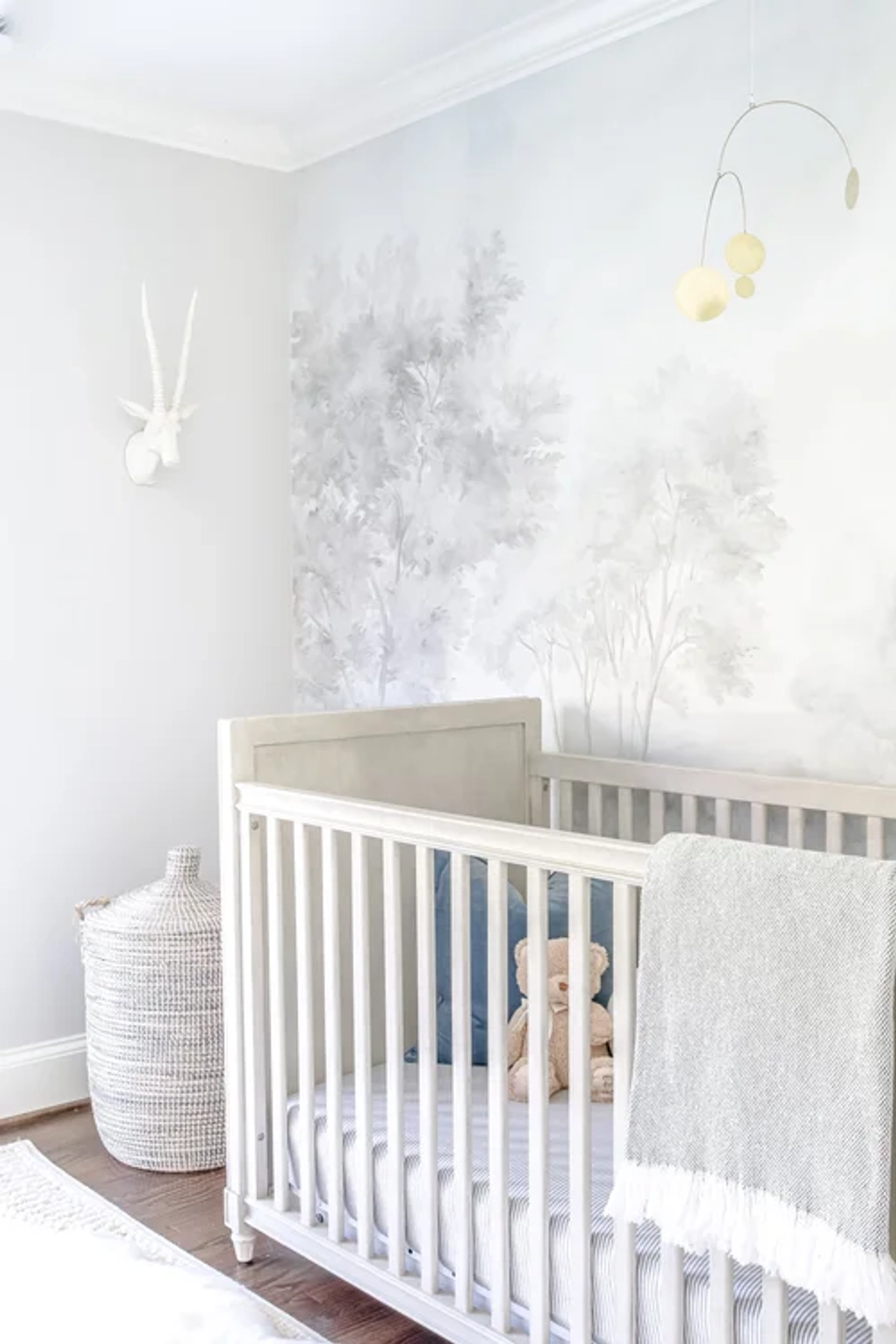 Close-up of a white wooden crib with striped bedding set against a soft gray watercolor tree mural wallpaper, with a hanging mobile and a white faux animal head wall mount