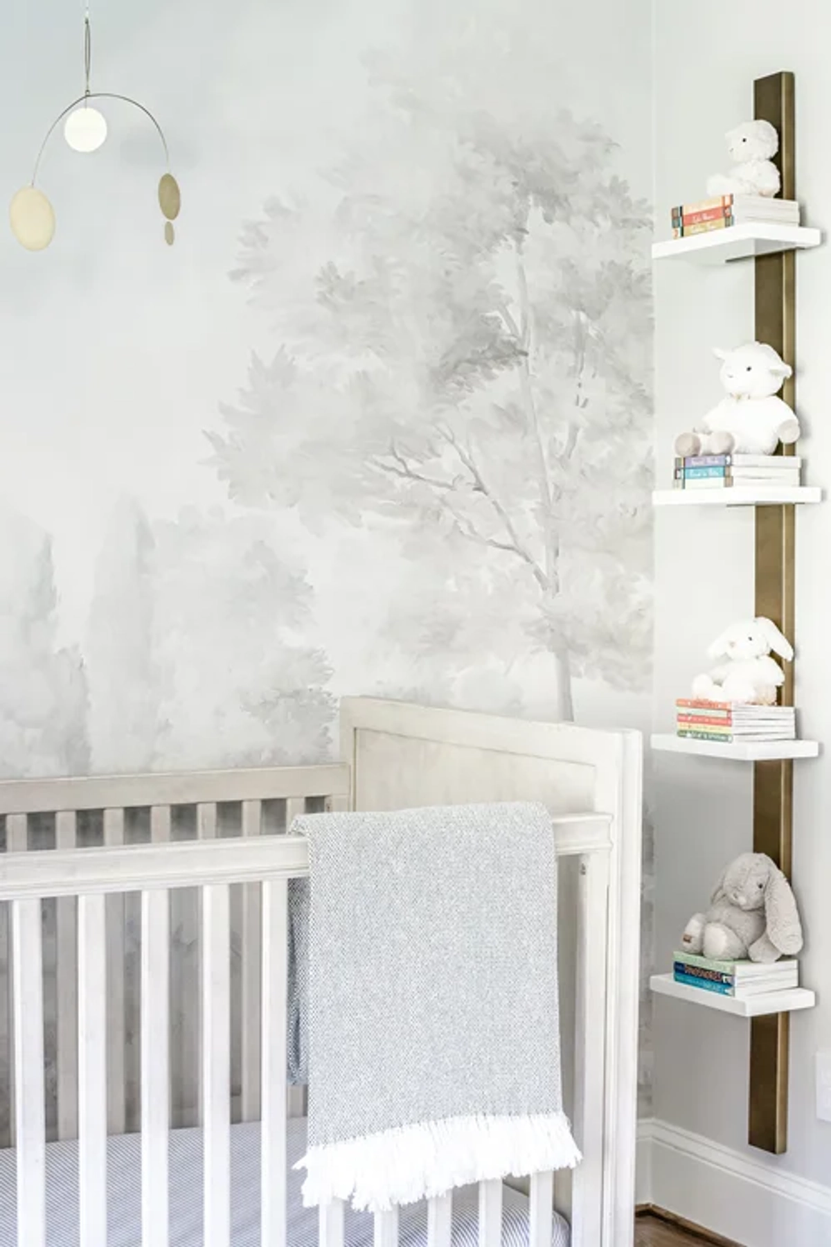 Nursery corner showing a white crib against gray tree mural wallpaper, a brass and wood mobile, and floating wall shelves with stuffed animals and children's books