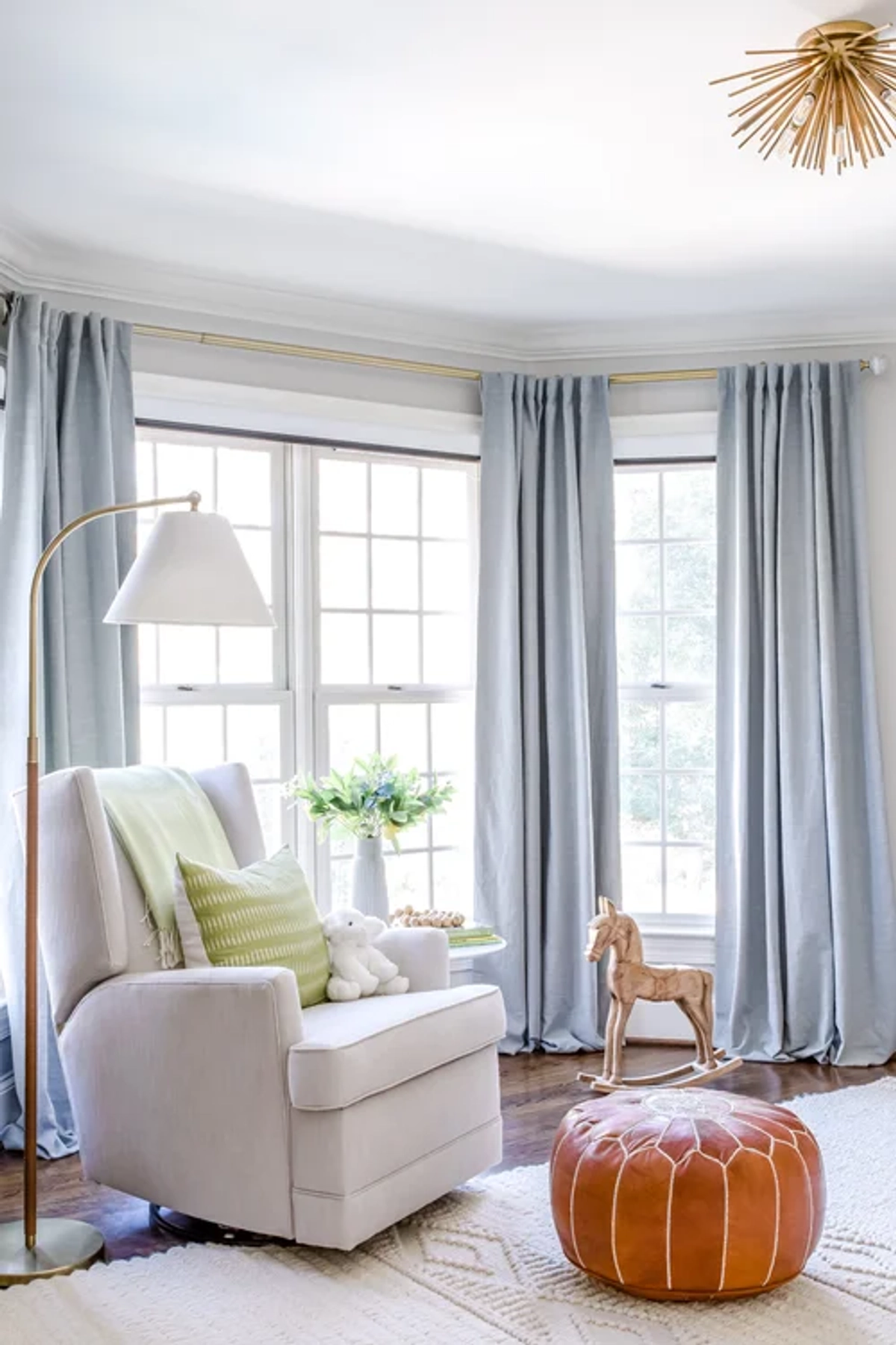 Nursery seating corner with a cream upholstered glider chair, a floor lamp, a cognac leather round pouf, soft blue curtains on a bay window, and a gold starburst ceiling light