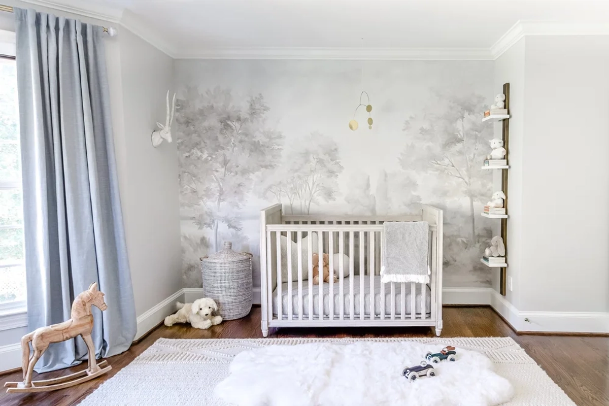 Full nursery view showing a white crib centered on a gray watercolor tree mural accent wall, light blue curtains, floating shelves with toys, and a wooden rocking horse