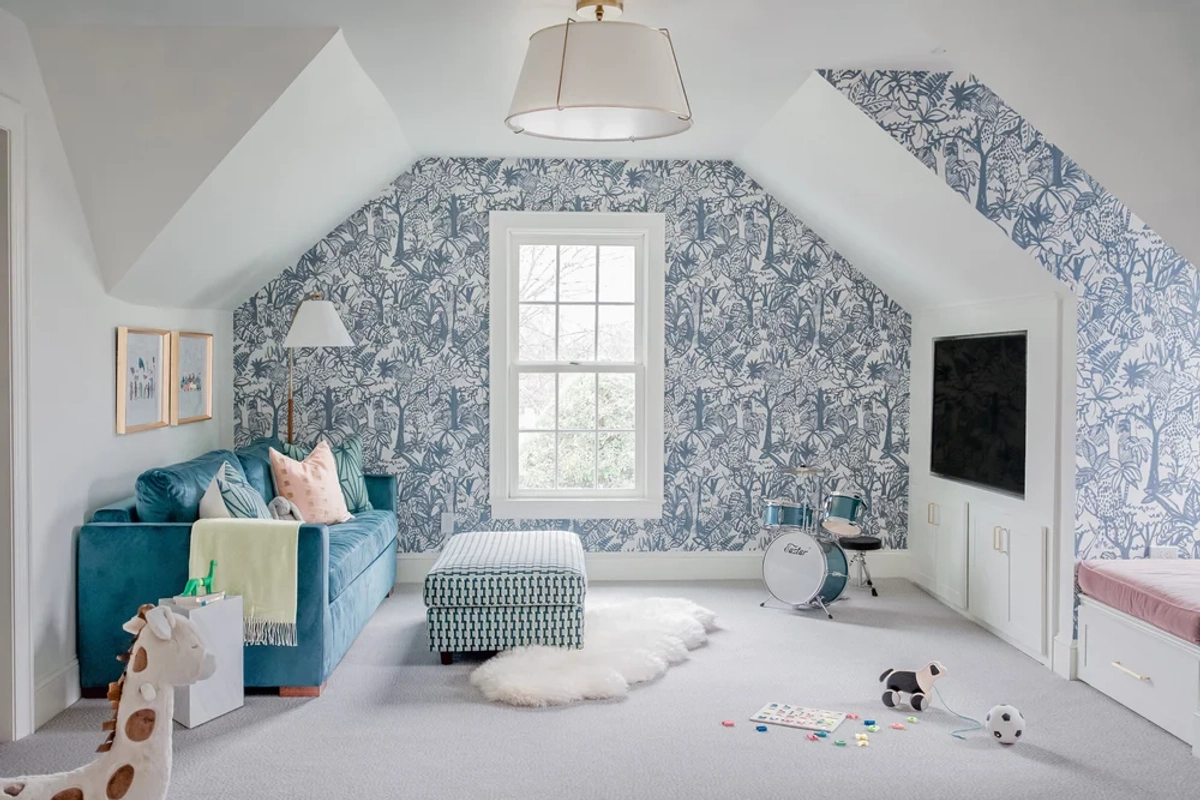 Attic playroom with blue-and-white jungle-themed toile wallpaper, a teal sofa with colorful pillows, a gingham ottoman, white built-in storage, and a drum pendant light