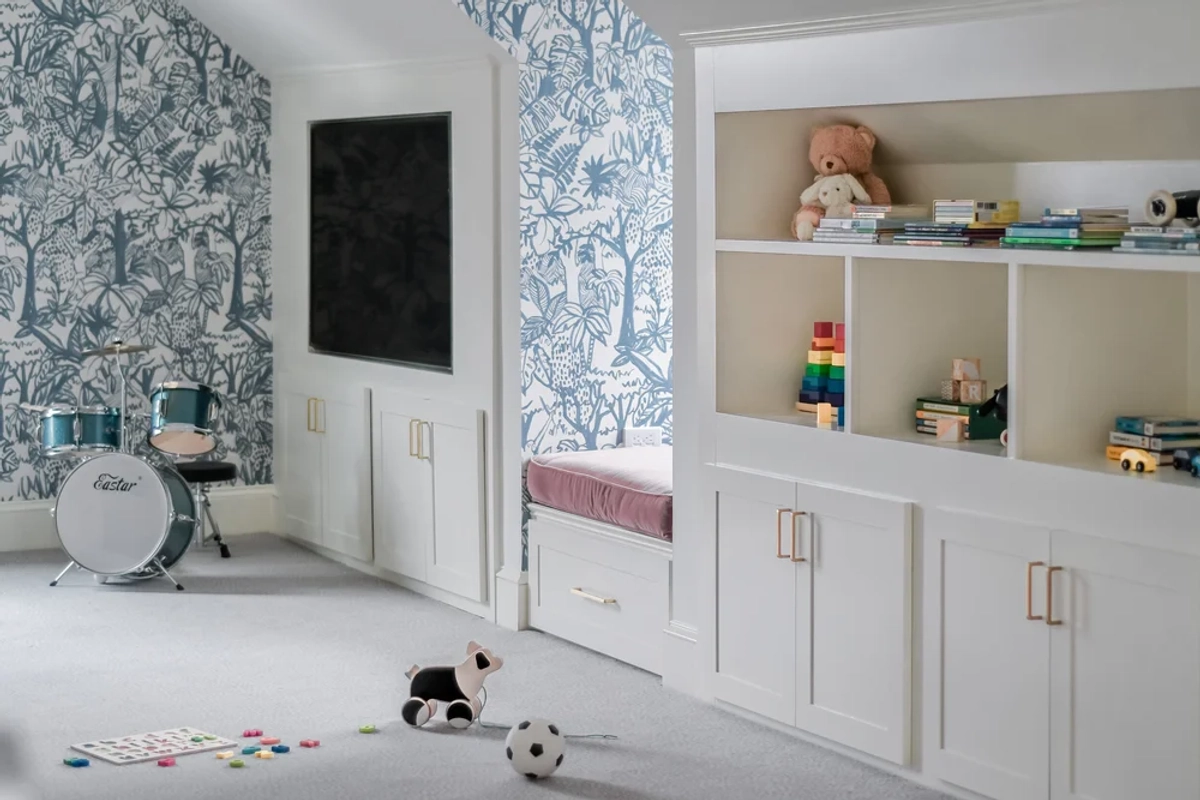Playroom wall featuring blue-and-white tropical wallpaper alongside white built-in cabinets with a window seat bench, brass hardware, and children's toys and books