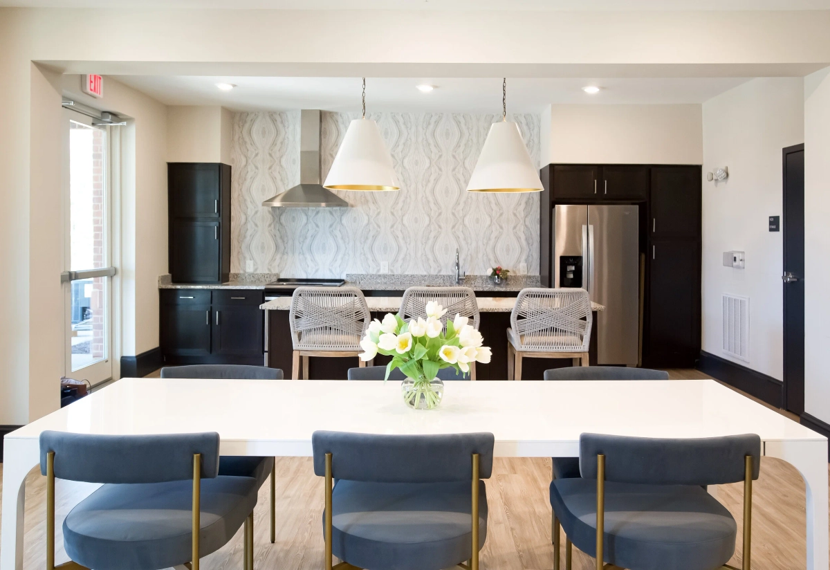 Community kitchen and dining area with dark cabinetry, stainless steel appliances, patterned wallpaper backsplash, white pendant lights, and a white dining table with navy blue upholstered chairs