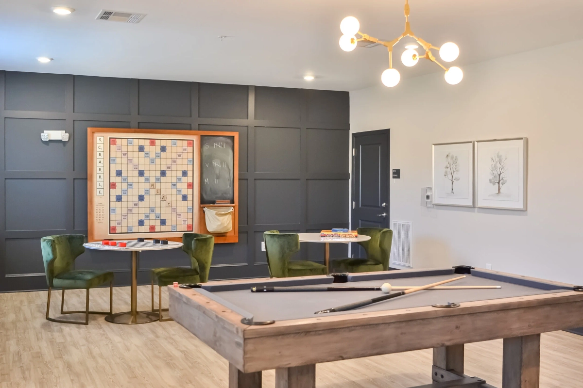Game room with a wood-framed pool table, oversized wall-mounted Scrabble board against a dark gray paneled wall, green velvet chairs at a small round table, and a gold sputnik chandelier