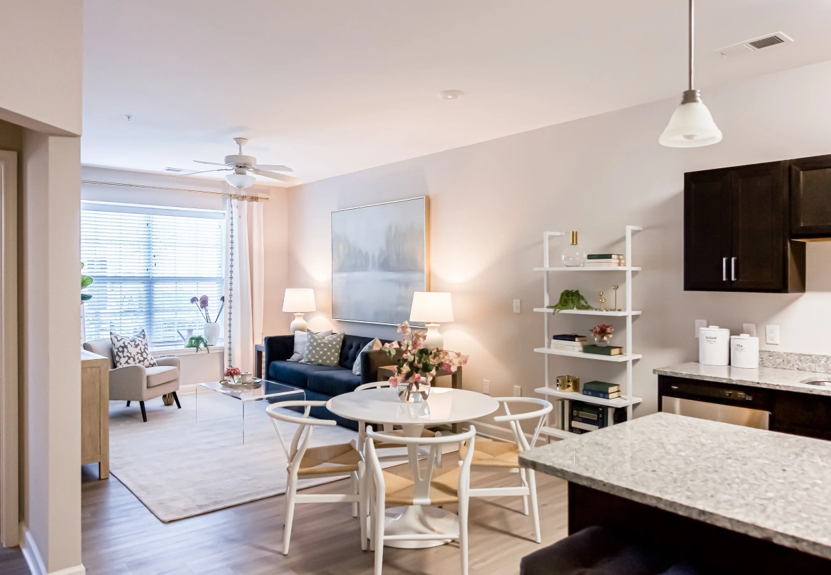 Open-concept model apartment with a navy blue sofa, white wishbone dining chairs around a round table, a white open bookshelf, granite kitchen countertop, and light hardwood flooring