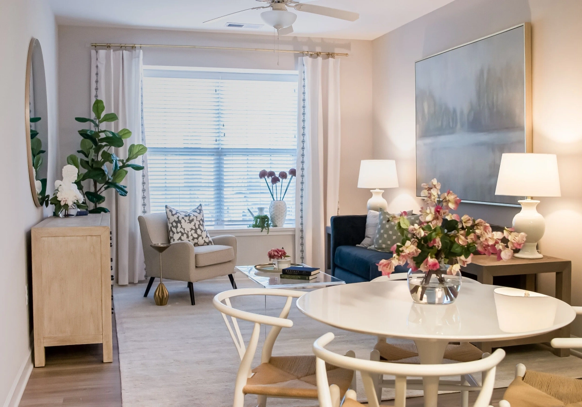 Bright living and dining area with a navy sofa, cream accent chair, white round dining table with wishbone chairs, a light wood console with round mirror, and fresh floral arrangements