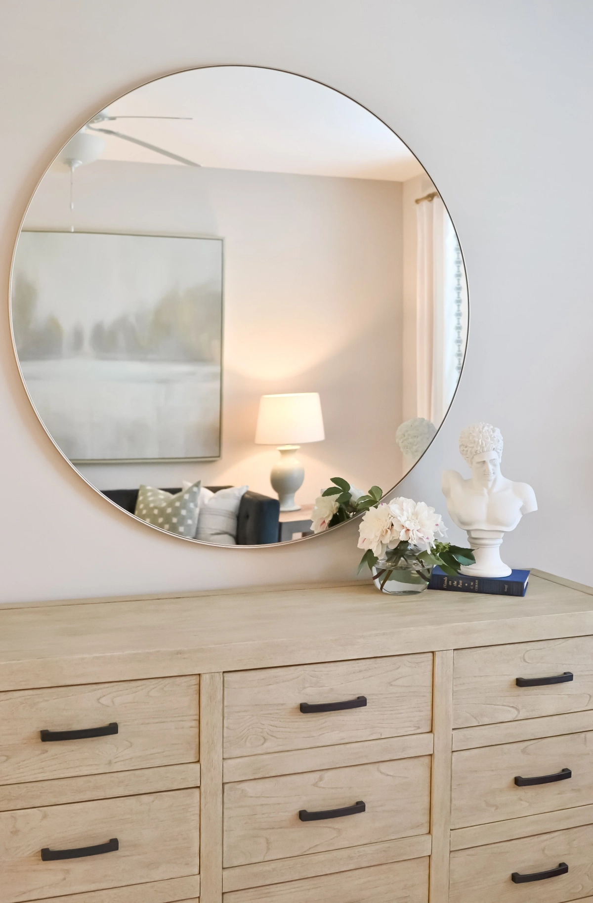Light oak dresser with black hardware topped by a large round mirror reflecting the living room, a white classical bust sculpture, white hydrangea arrangement, and stacked books