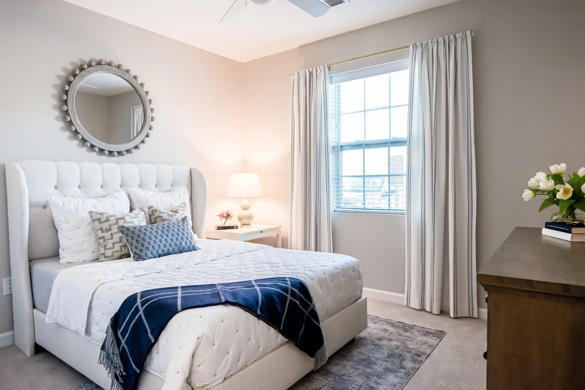 Model bedroom with a light gray tufted upholstered bed in white bedding and a navy plaid throw, decorative sunburst mirror above the headboard, bedside lamp, and fresh white tulips on a wood dresser