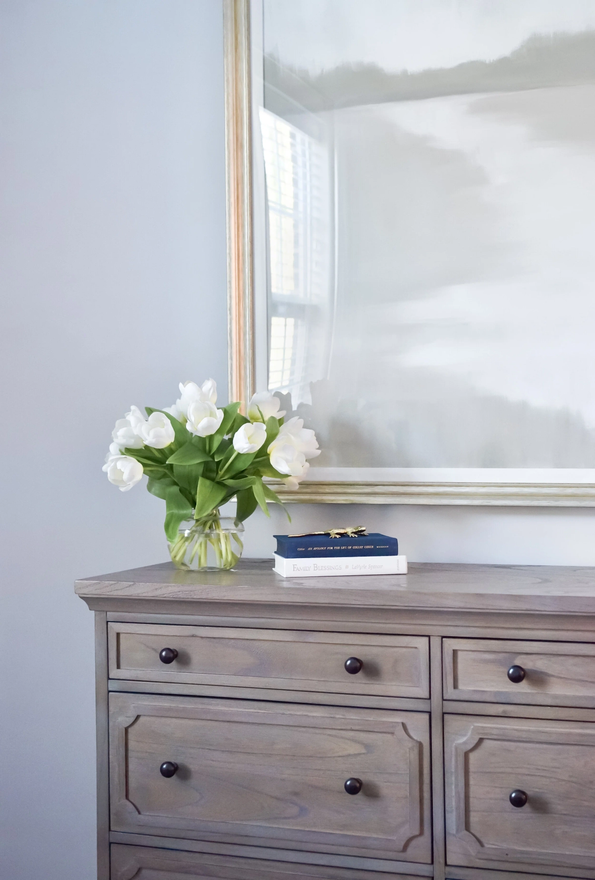 Weathered wood dresser with dark round knobs displaying a vase of white tulips and stacked books beneath a gold-framed abstract landscape painting against a soft blue wall
