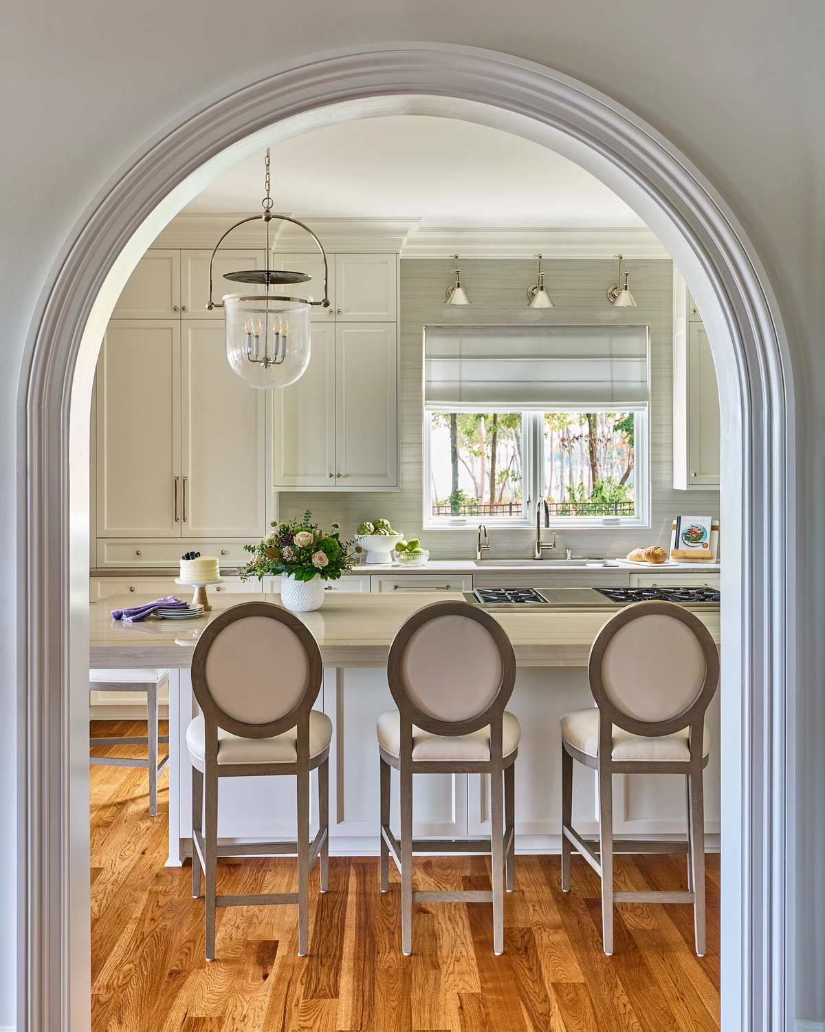 View through a white rounded archway into the kitchen with three gray oval-back bar stools at a marble island, a glass bell jar pendant, and white cabinetry