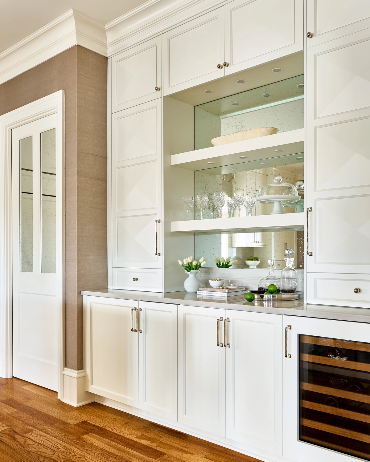 Built-in white bar cabinet with mirrored backsplash, open display shelves holding crystal glassware, a built-in wine refrigerator, polished nickel hardware, and grasscloth-covered adjacent wall