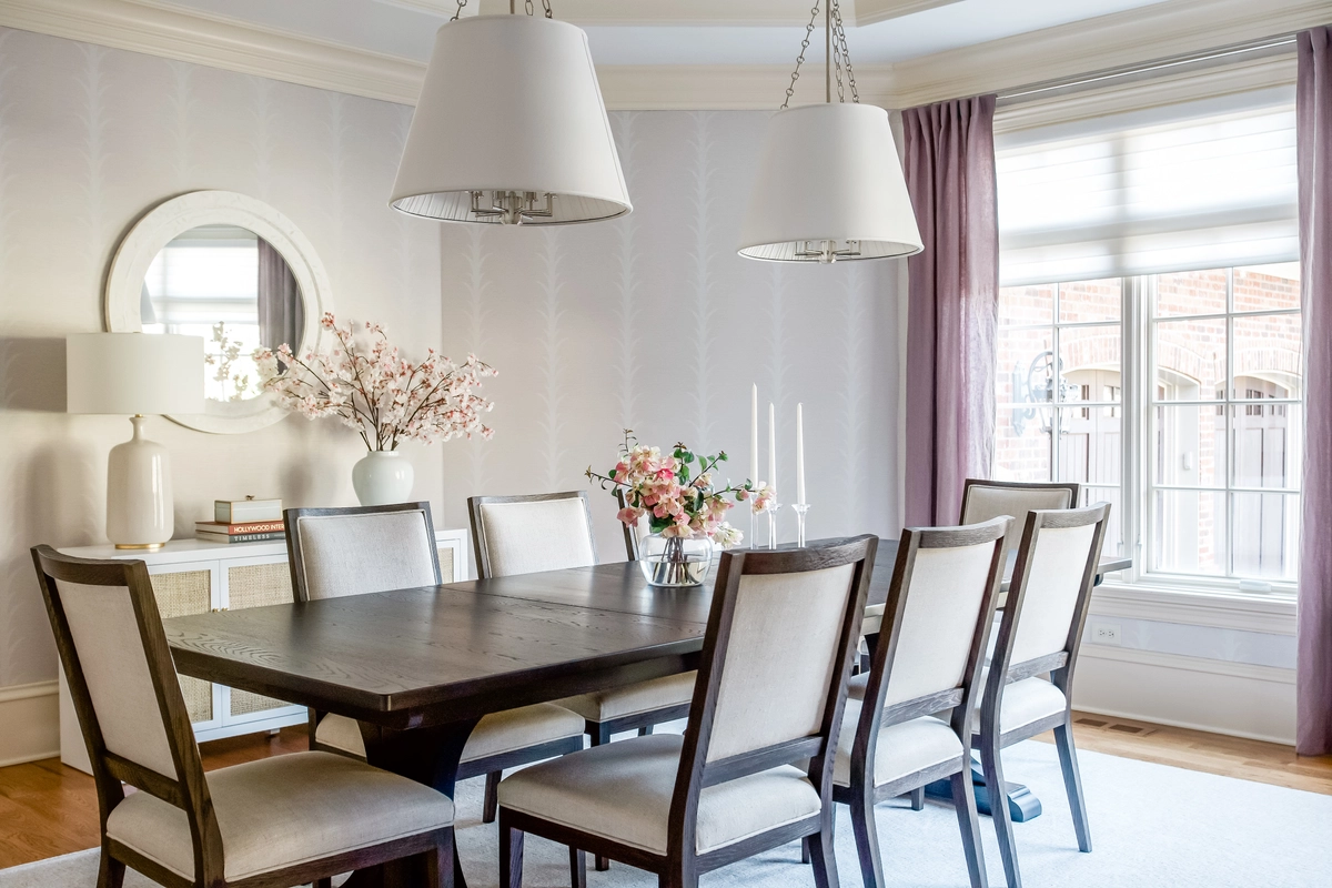 Dining room with a dark wood trestle table, cream upholstered chairs, dual drum pendant lights, mauve curtains, and a console with ceramic vases and cherry blossoms
