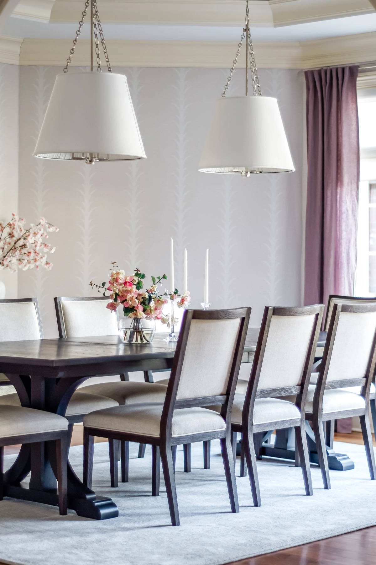 Close-up of the dining table with dual nickel drum pendants, fresh floral centerpiece, taper candles, and mauve linen curtains