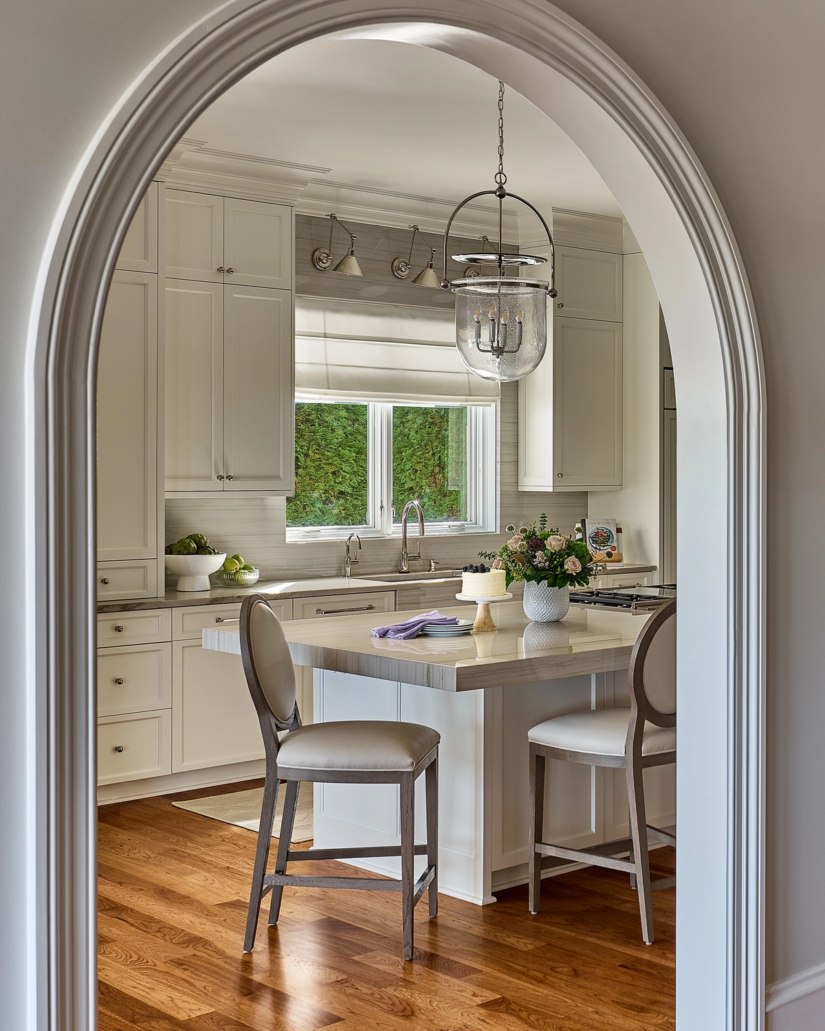 Portrait view through a white archway into the kitchen showing bar stools at the island, a glass pendant light, white floor-to-ceiling cabinetry, and a window overlooking greenery