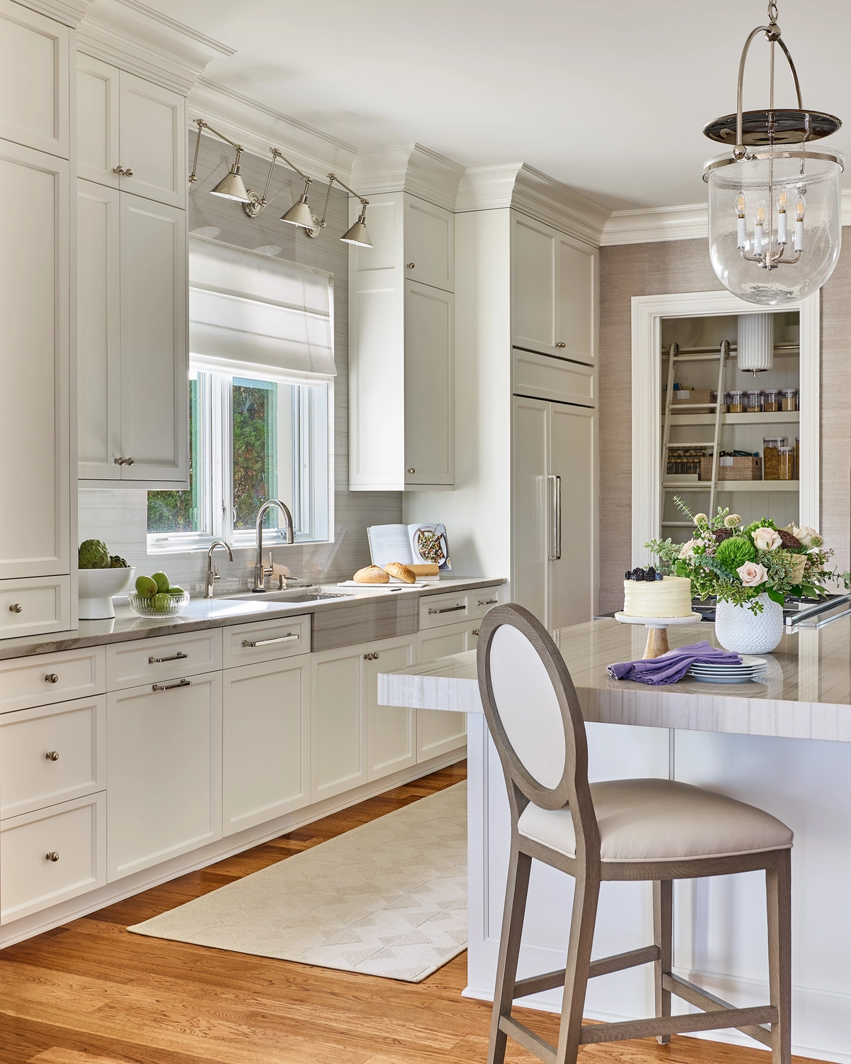 Kitchen angle showing white cabinetry with polished nickel pulls, a farmhouse sink beneath a window with roman shade, and an open doorway revealing an organized walk-in pantry with a rolling ladder