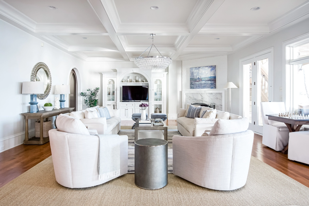 Bright living room with white swivel chairs, cream sofas, a crystal tiered chandelier, coffered ceiling, built-in arched cabinetry, and hardwood floors