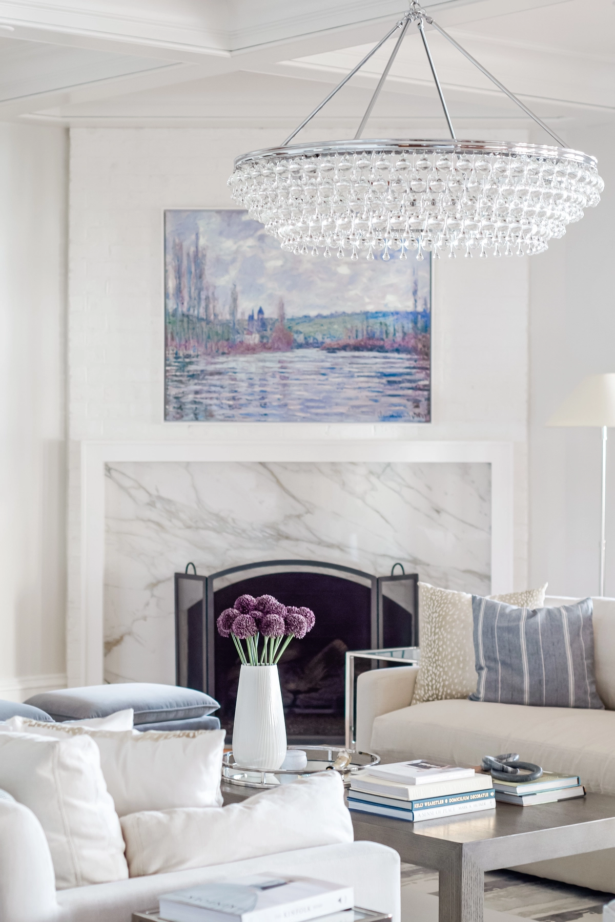 Living room fireplace with marble surround, tiered crystal chandelier, impressionist landscape painting, and cream upholstered seating with blue accents