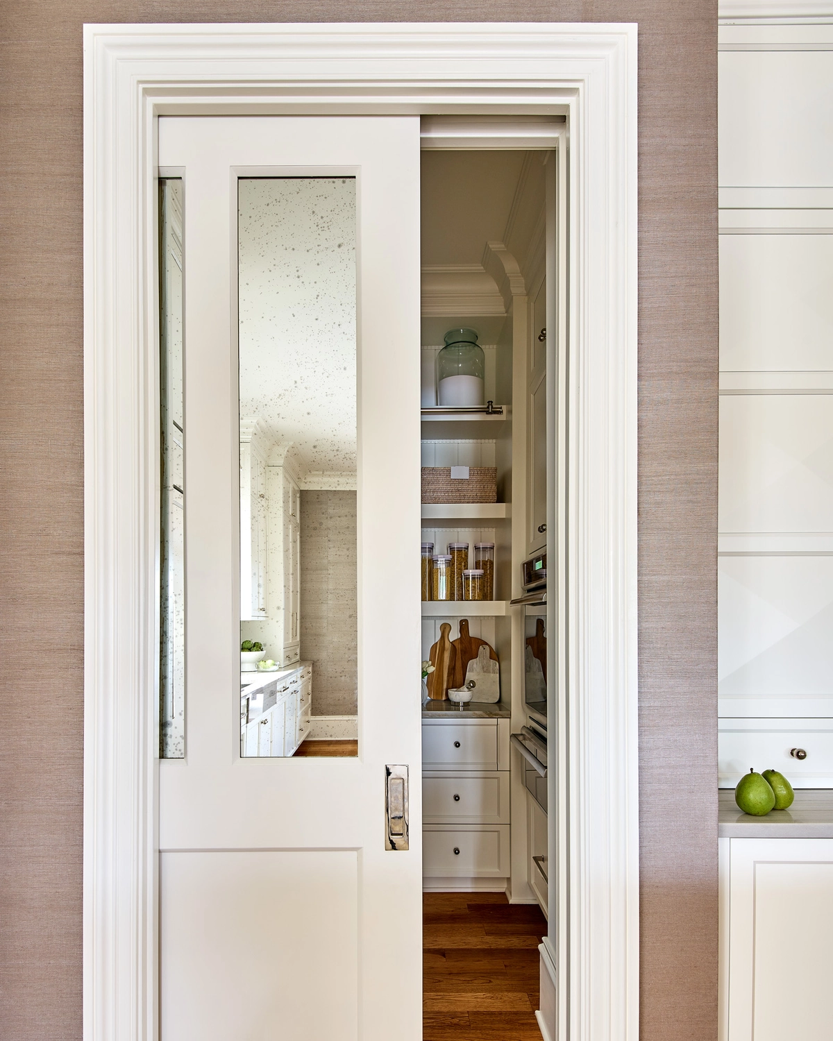 White pocket door with an antique mirrored glass panel partially open to reveal a walk-in pantry with built-in shelving, set against a taupe grasscloth wallpapered wall