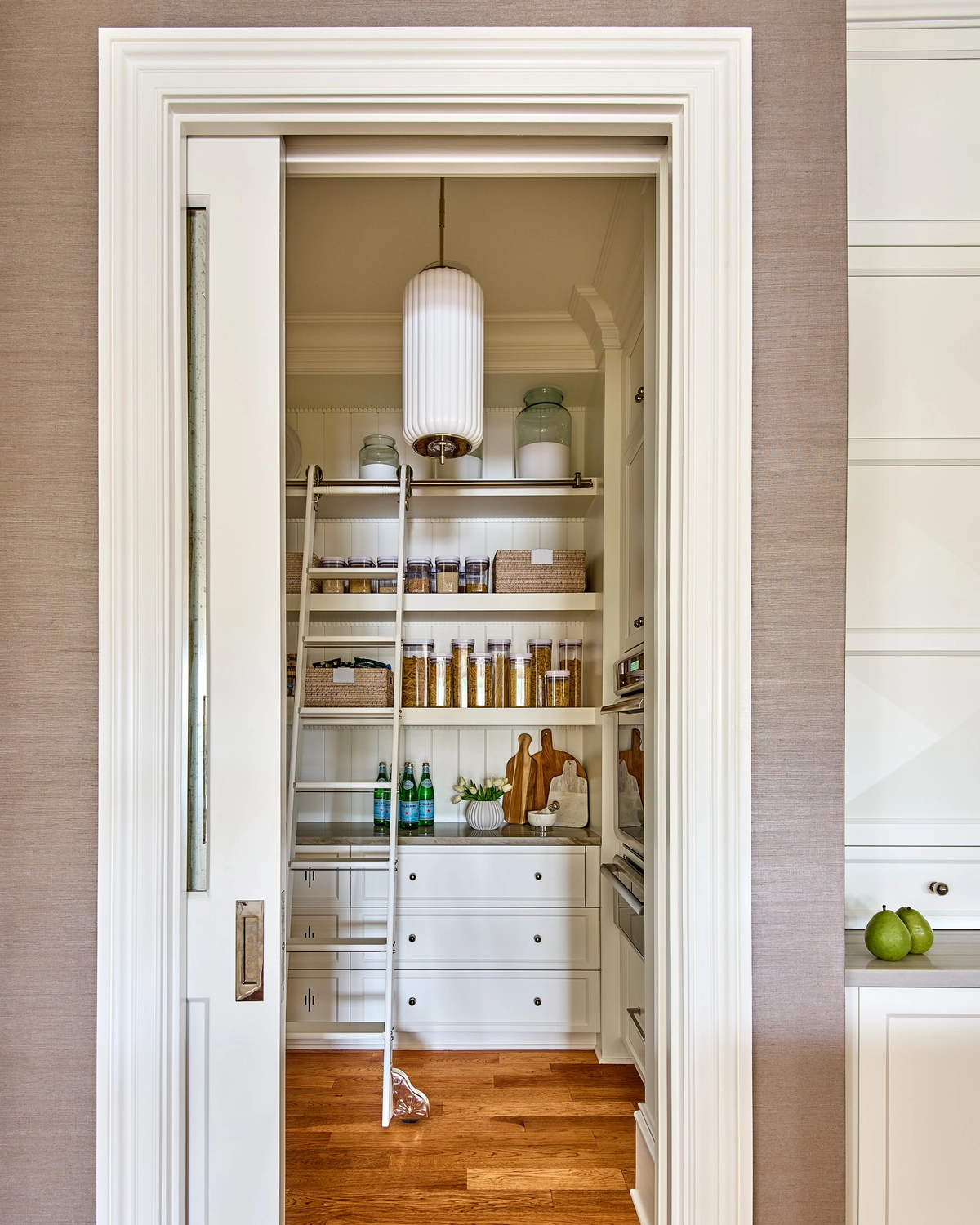 Walk-in pantry interior with a rolling nickel library ladder, open shelves stocked with glass jars and cookware, built-in white drawers, and a ribbed pendant light