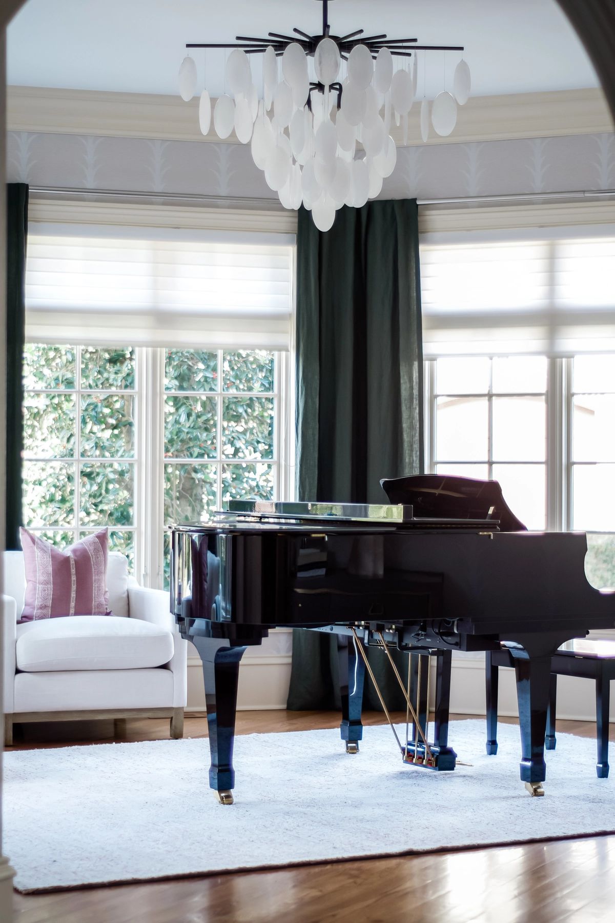Piano room viewed through an archway with a black grand piano, white disc chandelier, dark green curtains, and a white armchair with mauve pillow
