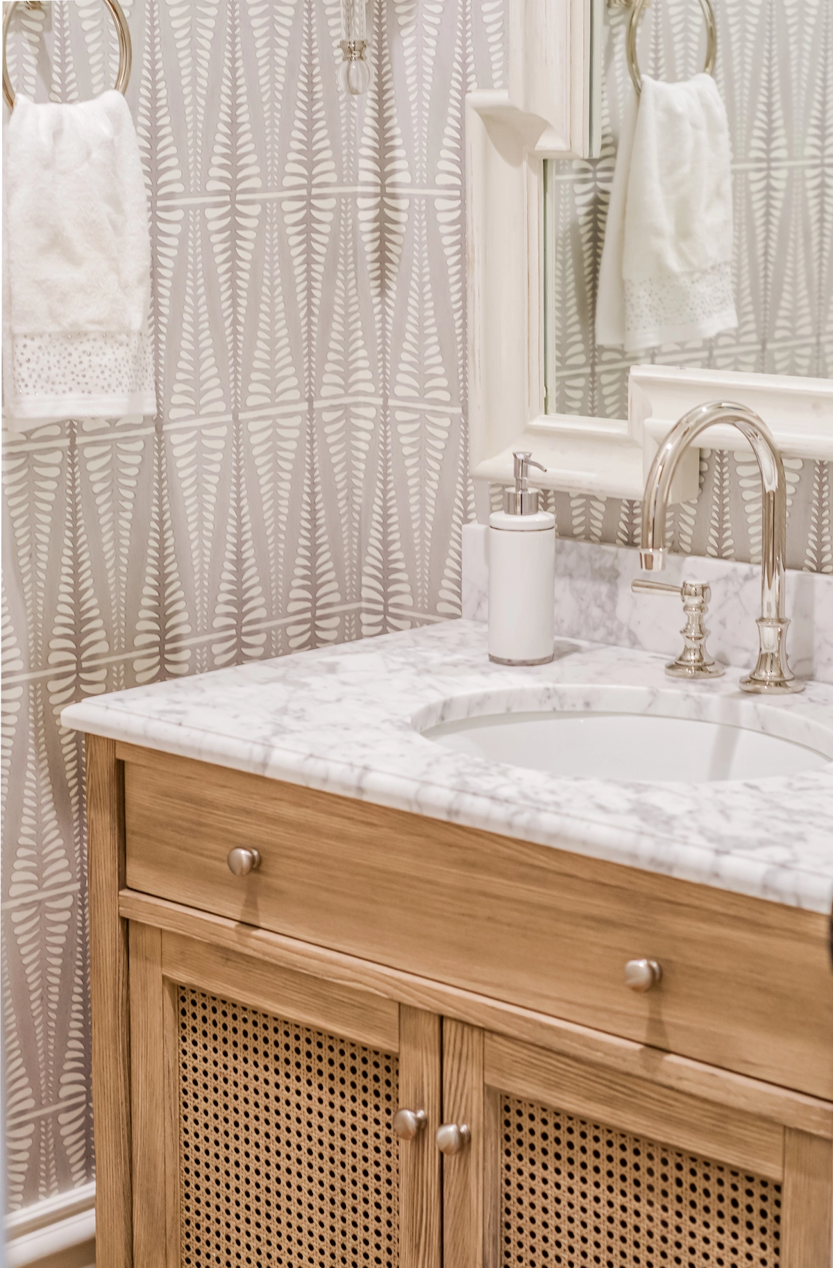 Powder bath with an oak and cane vanity, marble countertop, polished nickel faucet, fern-patterned wallpaper, and a framed mirror