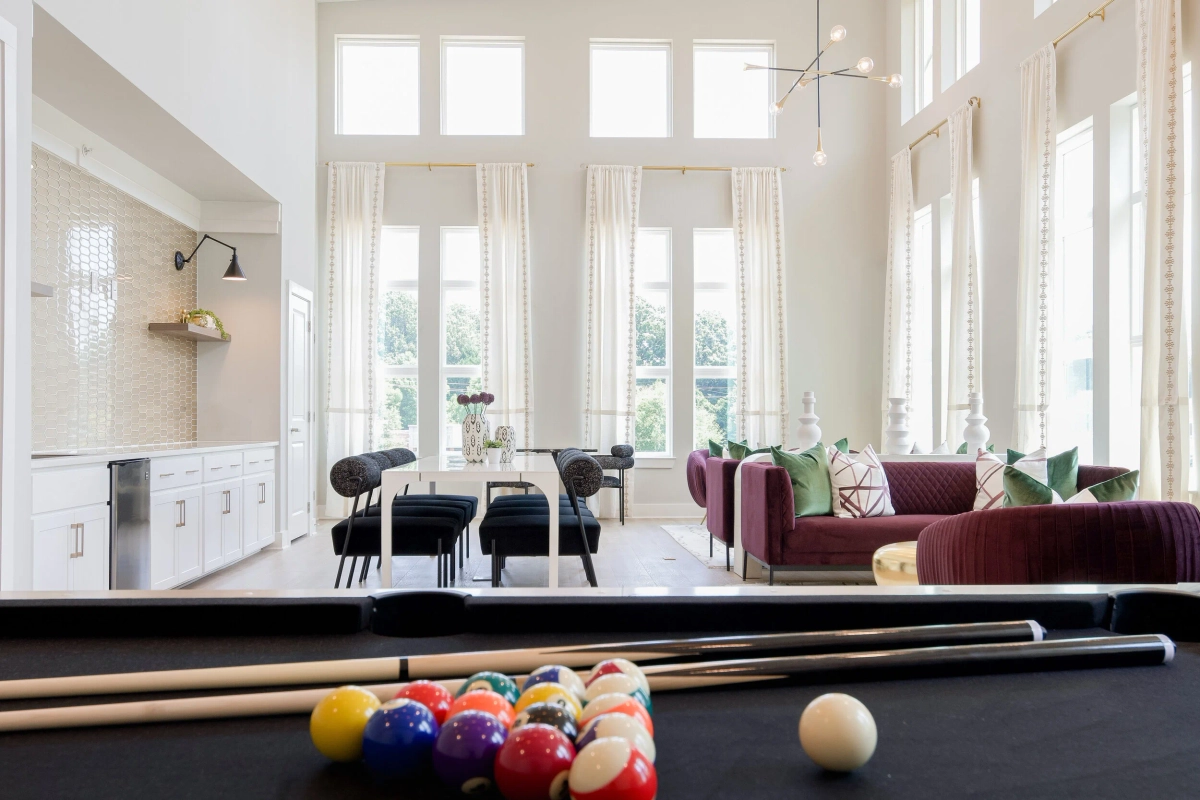 Double-height clubhouse lounge viewed from behind a black pool table, featuring floor-to-ceiling windows with sheer curtains, a plum velvet sofa, navy dining chairs, and a white kitchenette along the far wall