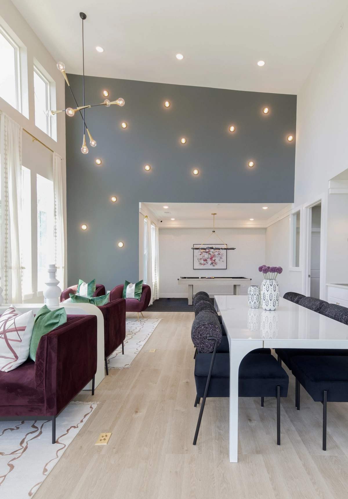 Clubhouse lounge with a dramatic dark teal accent wall adorned with scattered exposed-bulb lights, plum velvet sofas with green accent pillows, a white dining table with navy chairs, and a sputnik chandelier