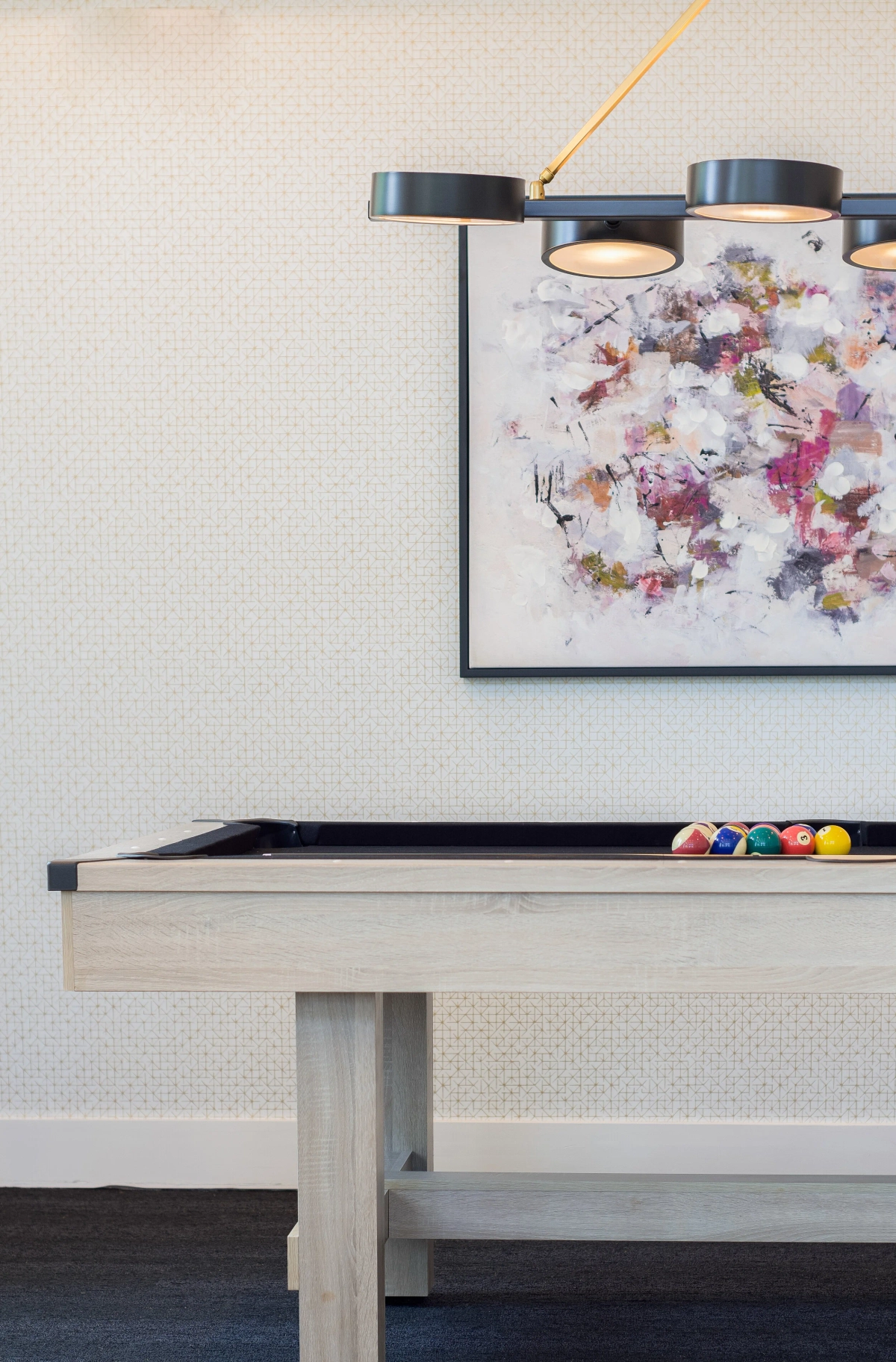 Light wood pool table with racked billiard balls beneath a modern matte black three-shade pendant light, with a large colorful abstract floral painting on the textured white wall behind
