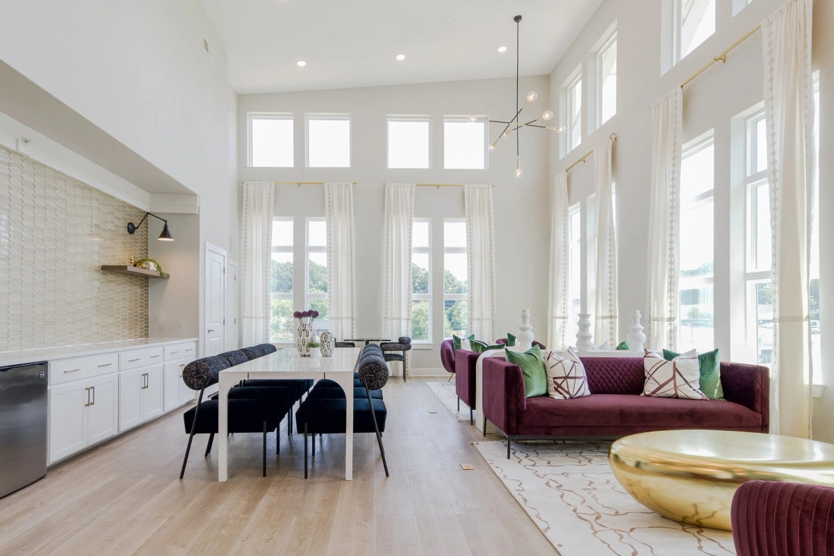 Wide view of the clubhouse showing a white built-in kitchenette with floating wood shelves and wall sconces, a white dining table with navy chairs, plum velvet sofas, and a gold drum coffee table