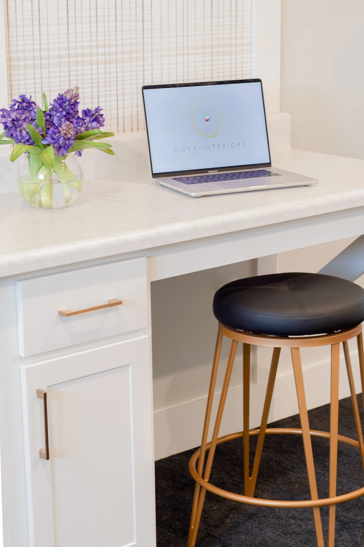 Built-in white desk nook with a quartz countertop, copper-toned drawer pulls, a laptop, a vase of purple hyacinths, and a black-and-gold wireframe stool