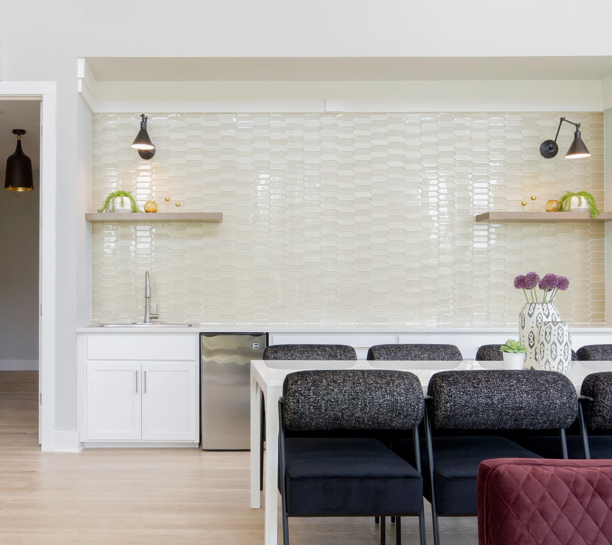 Clubhouse kitchenette featuring elongated hexagonal cream tile, floating wood shelves with decorative objects, matte black swing-arm sconces, white lower cabinets, and a stainless steel mini fridge