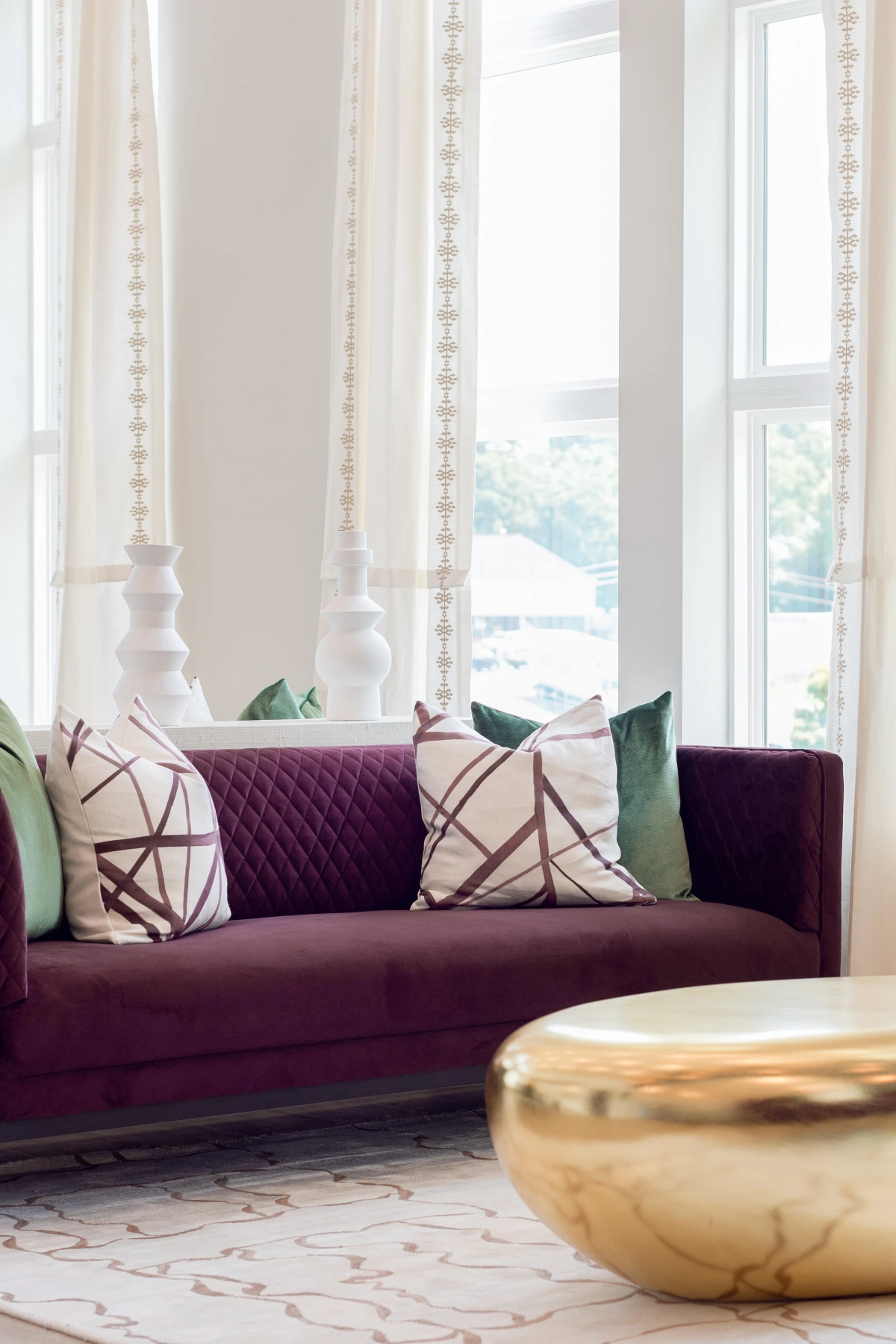 Deep plum quilted velvet sofa with green velvet and geometric patterned throw pillows, sheer pom-pom trimmed curtains against floor-to-ceiling windows, and a gold hammered drum coffee table