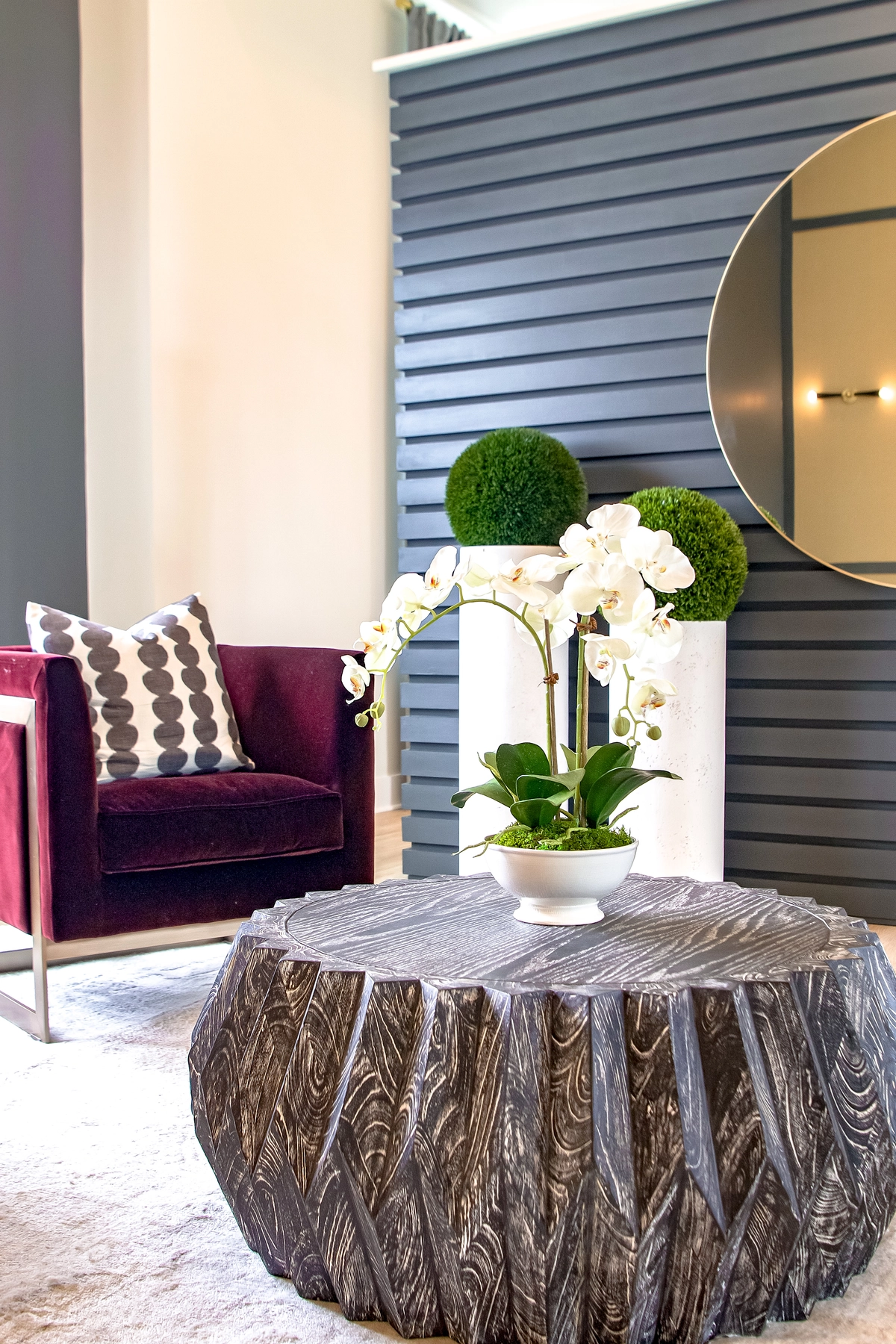 Lobby detail with a sculptural gray stone coffee table, white orchid arrangement, burgundy velvet armchair with geometric pillow, green moss topiaries, and a gray slatted accent wall with round mirror