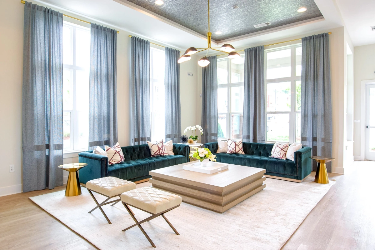 Elegant lobby lounge with two teal tufted velvet Chesterfield sofas, cream cross-leg stools, a stacked wood-and-white coffee table, sheer blue-gray curtains, a textured metallic ceiling, and a brass chandelier