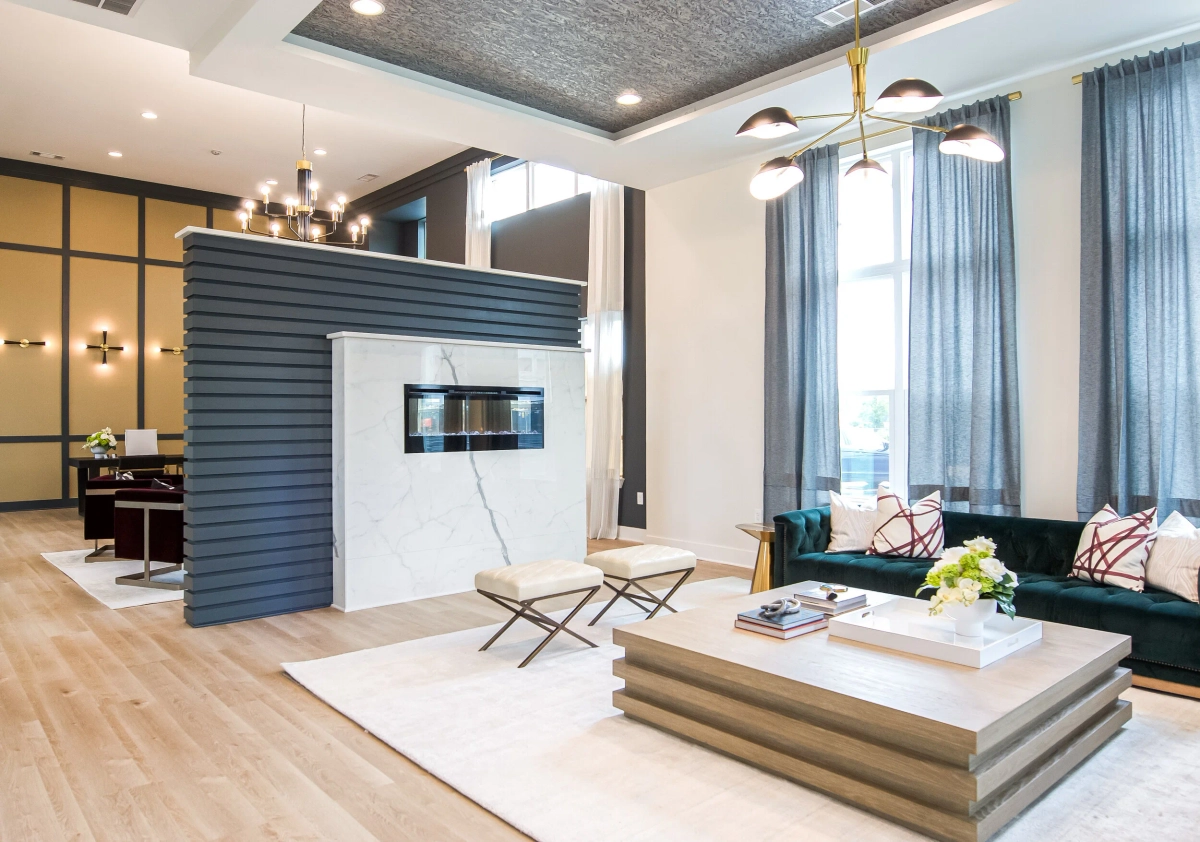 Open-plan lobby showing a gray slatted room divider with a marble-front linear fireplace, teal velvet sofas, brass pendant lighting, a textured ceiling, and a glass-walled office space