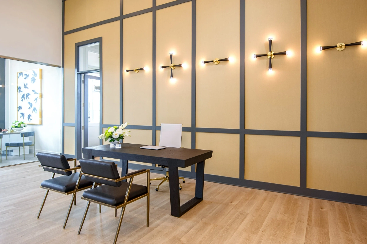 Leasing office with a dark wood desk and black leather chairs with brass legs, a mustard-gold paneled accent wall with gray trim and decorative starburst brass wall sconces