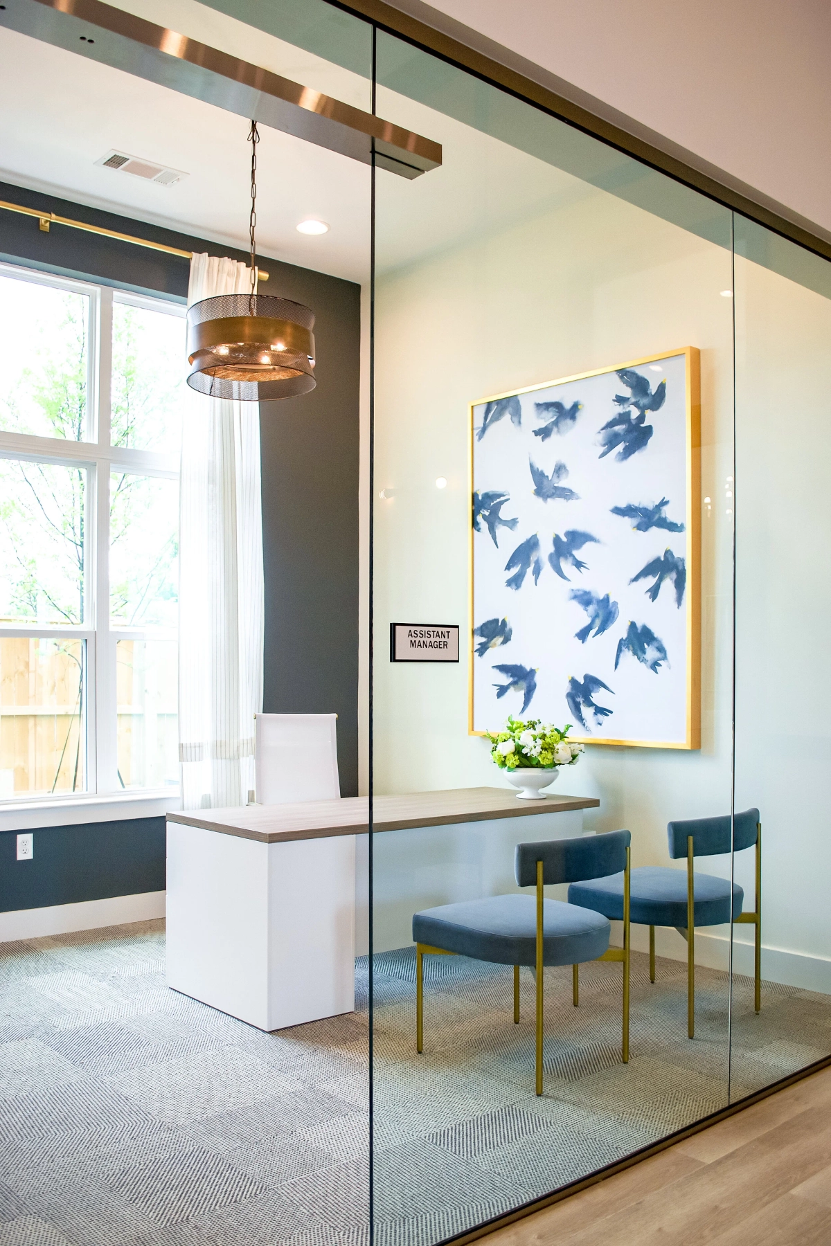 Glass-enclosed manager office with a white modern desk, blue upholstered guest chairs with brass legs, a copper drum pendant light, and a large blue bird painting in a gold frame