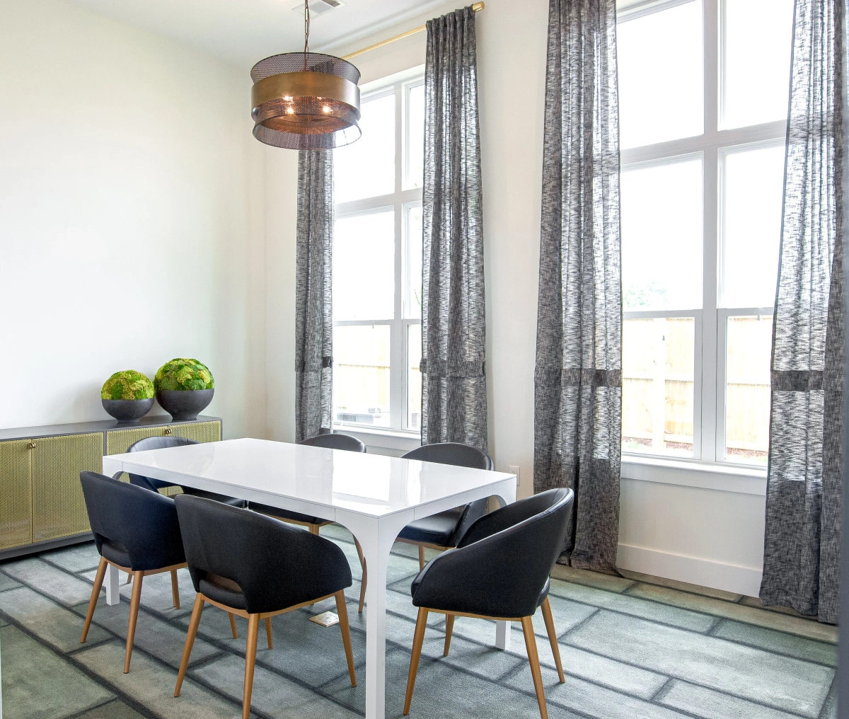 Conference room with a white table surrounded by dark charcoal mid-century chairs, a copper drum pendant light, gray patterned curtains, a gold credenza with green moss spheres, and a geometric area rug