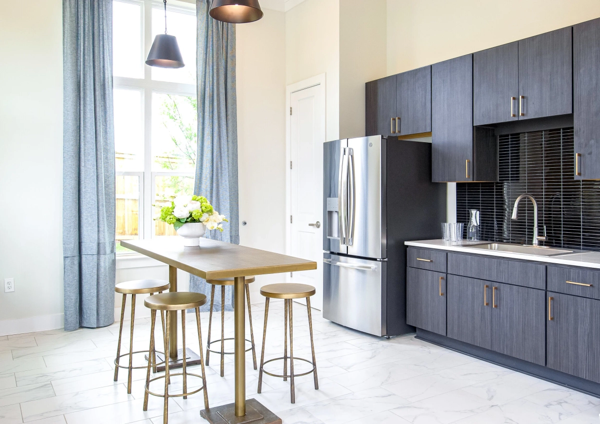 Community kitchen with dark gray cabinetry, stainless steel French-door refrigerator, black tile backsplash, a gold high-top table with brass bar stools, and marble-patterned tile flooring