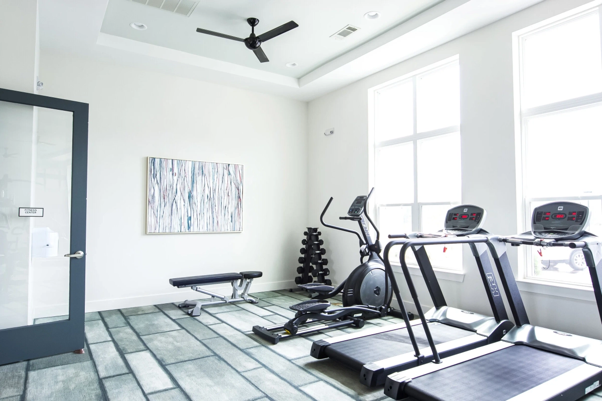 Fitness center with treadmills, an elliptical machine, a weight bench with dumbbell rack, abstract wall art, a ceiling fan, and large windows on gray stone-look flooring
