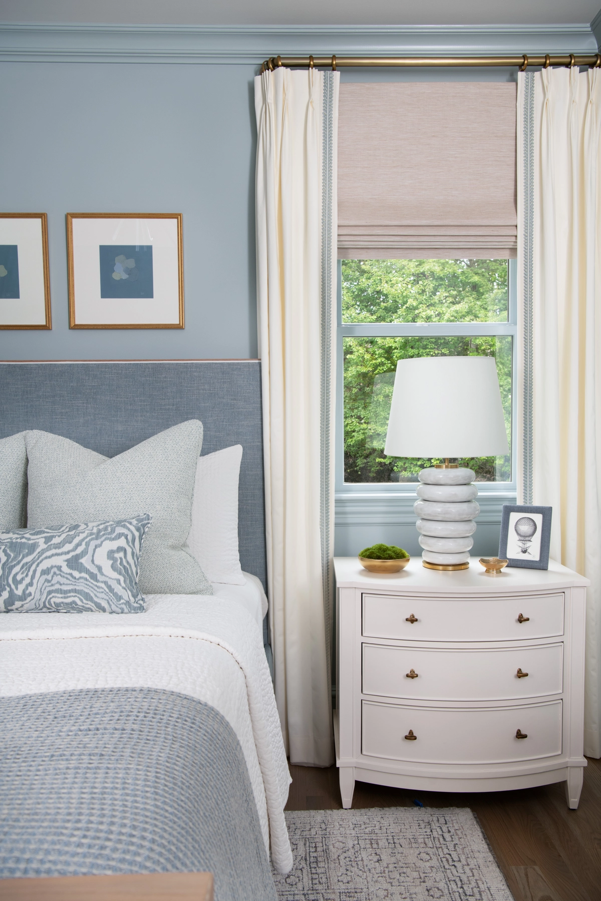 Soft blue bedroom corner with an upholstered blue headboard, white curved nightstand, stacked ceramic lamp, and ivory curtains framing a window with a woven Roman shade