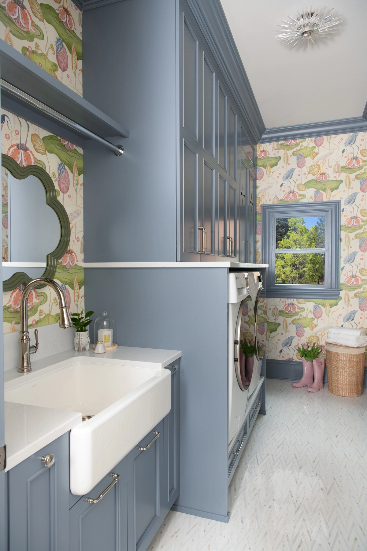 Charming laundry room with blue-gray cabinetry, a white farmhouse sink, colorful botanical wallpaper, a green quatrefoil mirror, and herringbone tile floors
