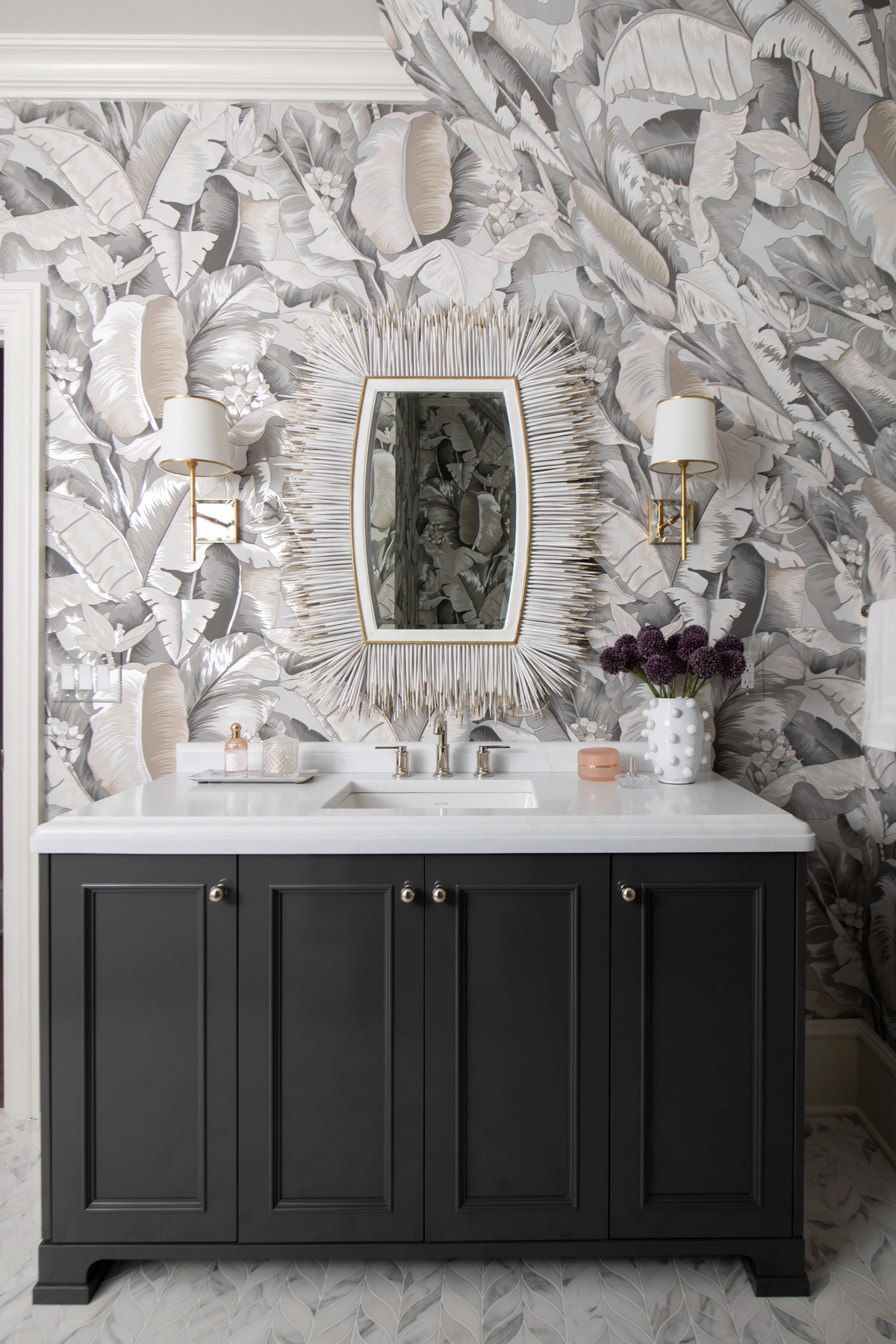 Powder room vanity with a black cabinet, white marble countertop, sunburst gold mirror, brass sconces, and dramatic gray botanical leaf wallpaper