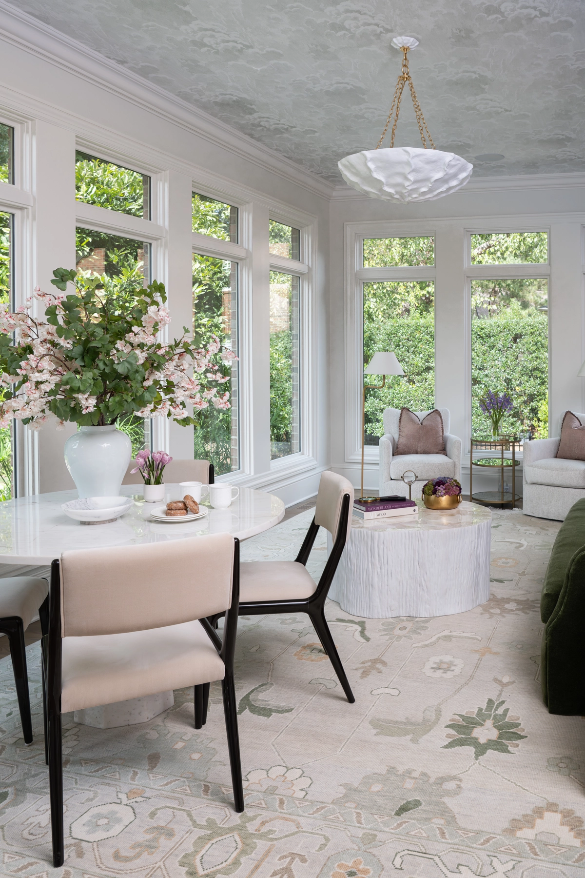 Light-filled sunroom with floor-to-ceiling windows, a marble dining table, cream upholstered chairs, olive green sofa, and a cloud-patterned ceiling with pendant light